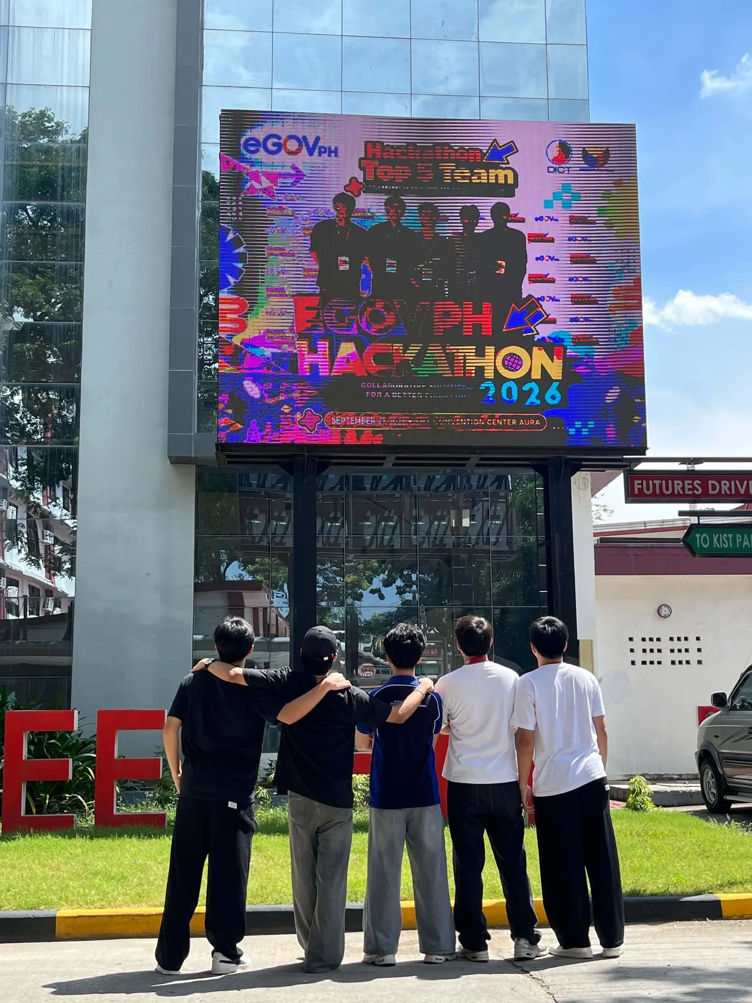 Five Theoraux team members seen from behind, arms around each other, looking up at an LED billboard showing the eGovPH Hackathon 2026 Top 5 Team announcement